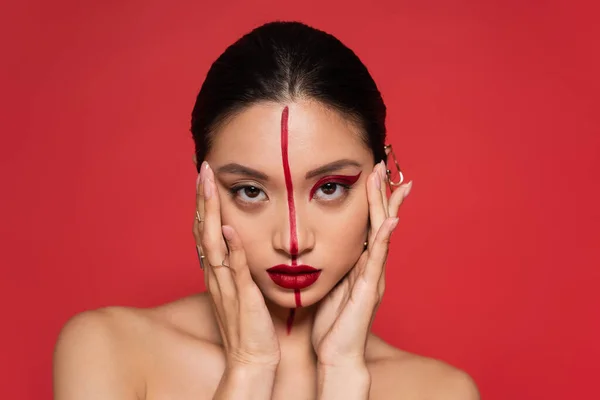 Brunette asian woman with bare shoulders and artistic makeup holding hands near face isolated on red — Stock Photo