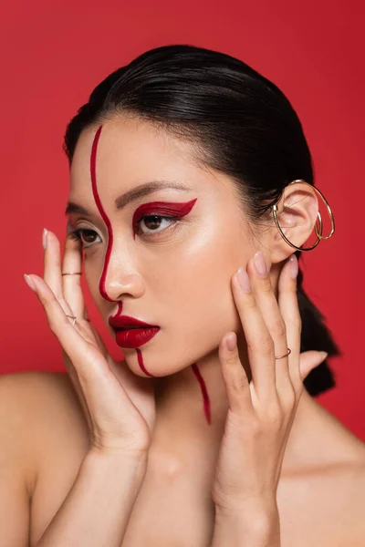 Portrait of asian woman with bright and creative makeup holding hands near face isolated on red — Stock Photo