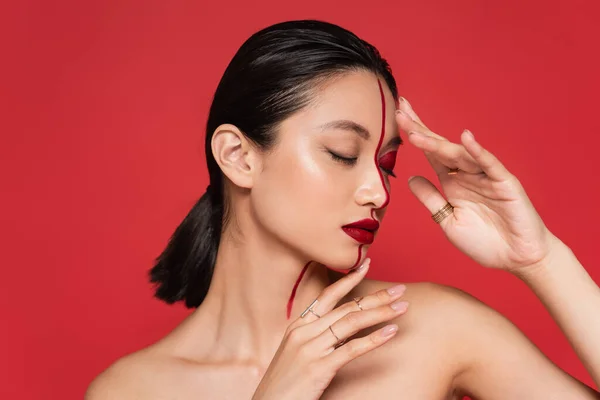 Sensual mujer asiática con hombros desnudos y ojos cerrados cogidos de la mano cerca de la cara con maquillaje creativo aislado en rojo - foto de stock