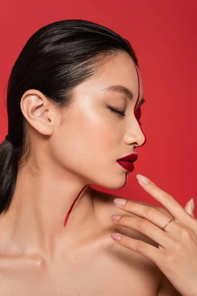 Perfil de mujer asiática con los ojos cerrados y rostro creativo sosteniendo la mano cerca de la cara aislada en rojo - foto de stock