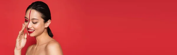 Щаслива азіатська жінка з закритими очима торкається обличчя з креативним макіяжем ізольовано на червоному, банер — стокове фото