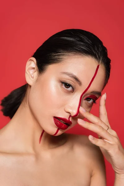 Porträt einer sinnlichen asiatischen Frau mit brillantem künstlerischem Make-up, die in die Kamera blickt und das Gesicht berührt, isoliert auf Rot — Stockfoto