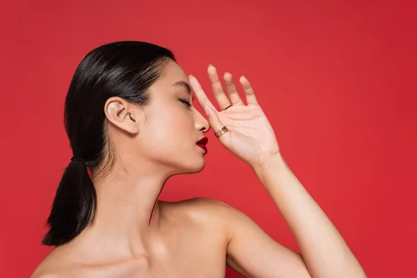 Perfil de morena mujer asiática con hombros desnudos y maquillaje posando con la mano cerca de la cara aislada en rojo - foto de stock