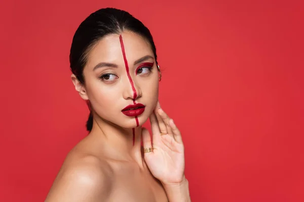 Jolie asiatique femme avec nu épaule et artistique visage touchant visage tout en regardant loin isolé sur rouge — Photo de stock