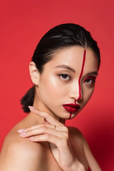 Junge asiatische Frau mit künstlerischem Aussehen blickt in die Kamera und berührt die nackte Schulter isoliert auf rot — Stockfoto