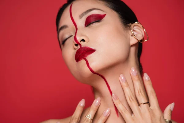 Спокуслива азіатська жінка з вушним манжетом і творчим макіяжем позує із закритими очима і руками біля шиї ізольовано на червоному — стокове фото