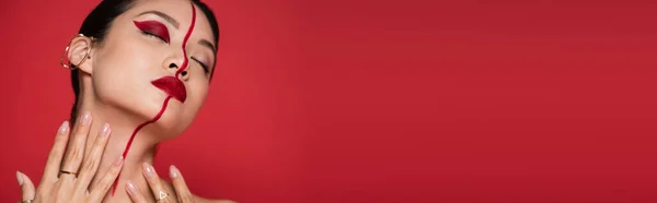 Sinnliche asiatische Frau in Ohrenmanschette und kreativer Visage, die den Hals berührt, während sie mit geschlossenen Augen auf rotem Banner posiert — Stockfoto