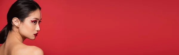 Perfil de morena asiática mulher no manguito da orelha e visagem criativa isolado no vermelho, banner — Fotografia de Stock
