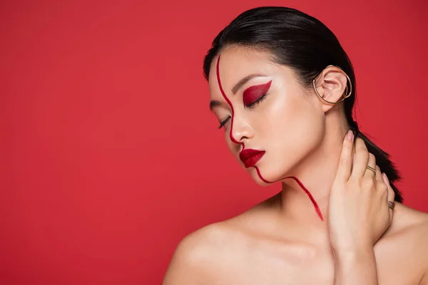 Seductive asian woman with bare shoulders and artistic makeup posing with closed eyes and hand on neck isolated on red — Stock Photo