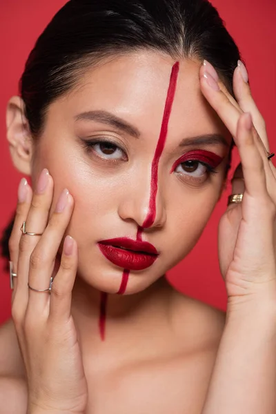 Портрет брюнетки азіатки дивиться на камеру, торкаючись обличчя з креативним візитом ізольовані на червоному — стокове фото