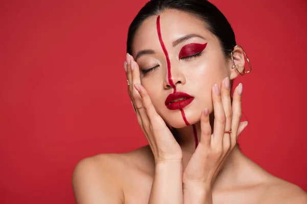 Seductive asian woman with bare shoulders and artistic visage touching face and posing with closed eyes isolated on red — Stock Photo