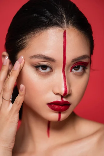 Porträt einer asiatischen Frau mit artistischem Make-up, die Gesicht berührt und isoliert auf rot in die Kamera blickt — Stockfoto