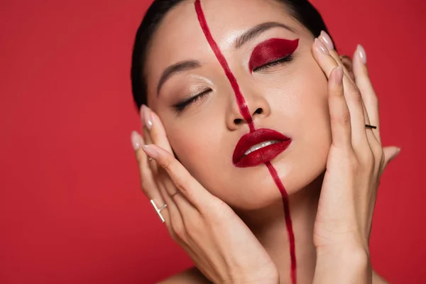 Retrato de sensual mujer asiática tocando perfecta cara con brillante maquillaje artístico mientras posando con los ojos cerrados aislados en rojo - foto de stock