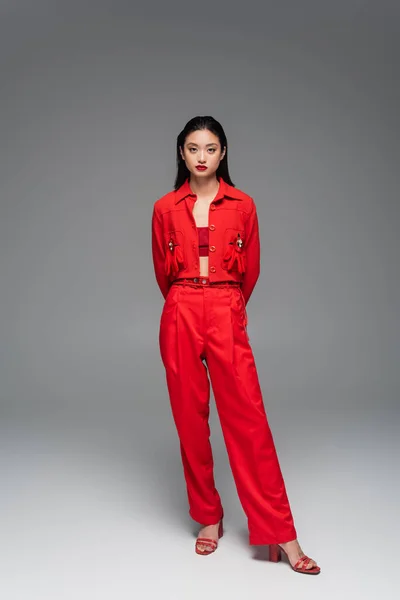 Longitud completa de la mujer asiática de moda en chaqueta roja y pantalones posando con las manos detrás sobre fondo gris - foto de stock