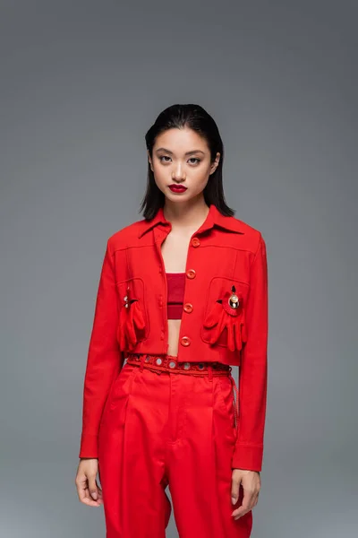 Молодая азиатка в красной куртке, украшенной брошками и перчатками, смотрящая на камеру, изолированную на сером — стоковое фото