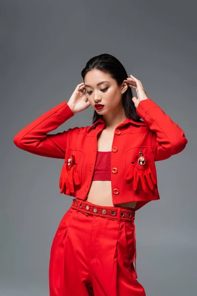 Brunette asian model in red jacket decorated with brooches and gloves fixing hair isolated on grey — Stock Photo