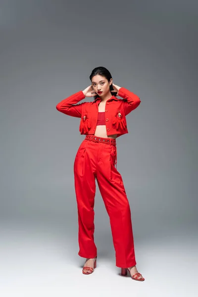 Повна довжина азіатської жінки в червоному елегантному вбранні позує руками за головою на сірому фоні — стокове фото