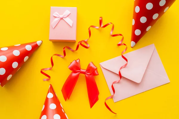 Top view of gift box near envelope and party caps on yellow background — Stock Photo