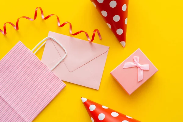 Top view of pink envelope and shopping bag near party caps and gift on yellow background — Stock Photo