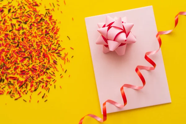 Top view of pink greeting card with gift bow and blurred sprinkles on yellow background — Stock Photo