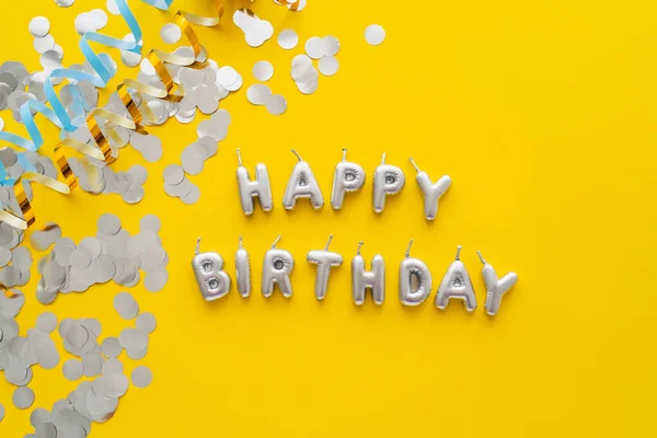 Vista superior de las velas en forma de letras de Feliz Cumpleaños y confeti sobre fondo amarillo - foto de stock