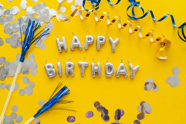 Top view of candles in shape of Happy Birthday lettering near confetti and serpentine on yellow background — Stock Photo