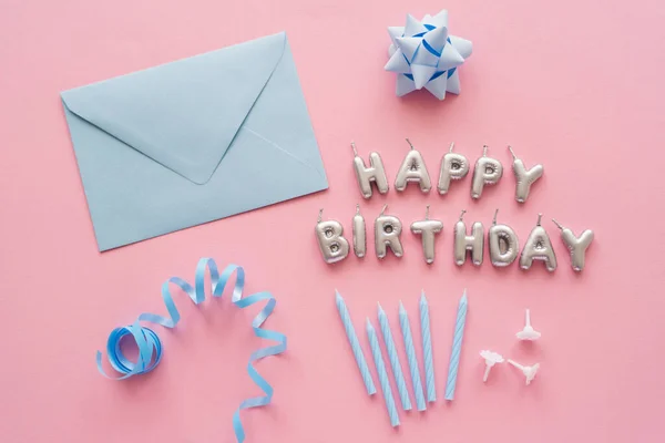 Ansicht von Kerzen in Form von Happy Birthday Schriftzug in der Nähe von blauem Umschlag und Serpentin auf rosa Hintergrund — Stockfoto