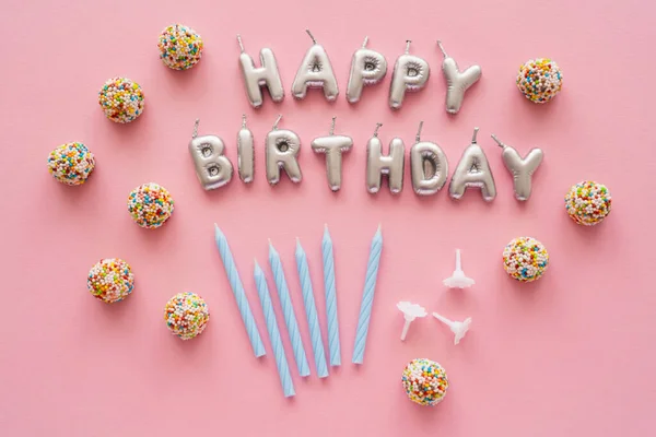 Vista superior de las velas en forma de letras de Feliz Cumpleaños cerca de caramelos sobre fondo rosa - foto de stock