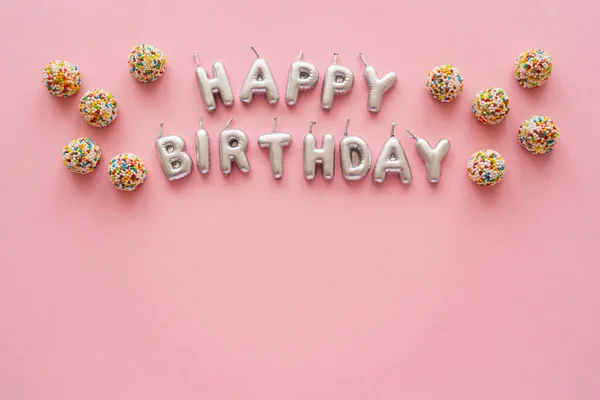 Vista superior de las velas en forma de letras de Feliz Cumpleaños cerca de dulces de colores sobre fondo rosa - foto de stock