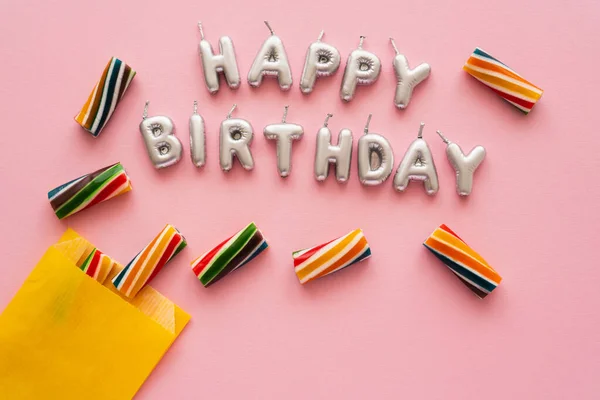 Top view of candles in shape of Happy Birthday lettering near striped sweets on pink background — Stock Photo