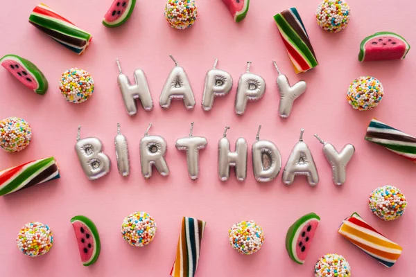 Vista superior de caramelos de colores cerca de velas en forma de letras de Feliz Cumpleaños sobre fondo rosa - foto de stock