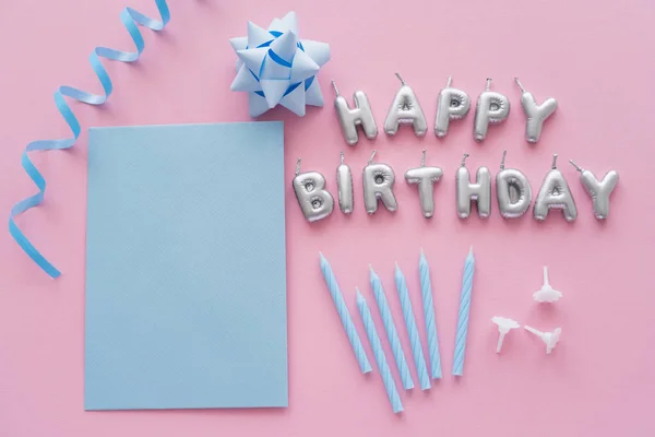 Top view of empty greeting card and candles in shape of Happy Birthday lettering near gift bow on pink background — Stock Photo
