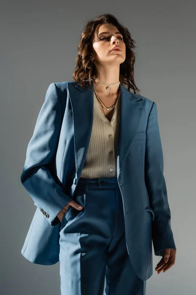 Уверенная в себе женщина в модном пиджаке, стоящая с рукой в кармане синих брюк, изолированных на сером — стоковое фото