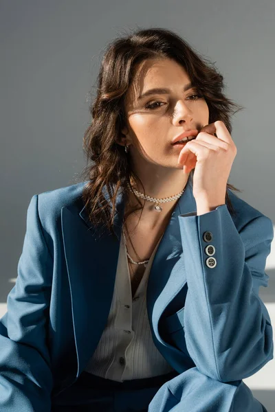 Thoughtful brunette woman in blue jacket and necklaces holding hand near face on grey background — Stock Photo