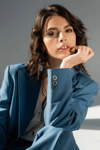 Портрет красивой брюнетки модели в синей и стильной куртке, смотрящей в камеру на сером фоне — стоковое фото