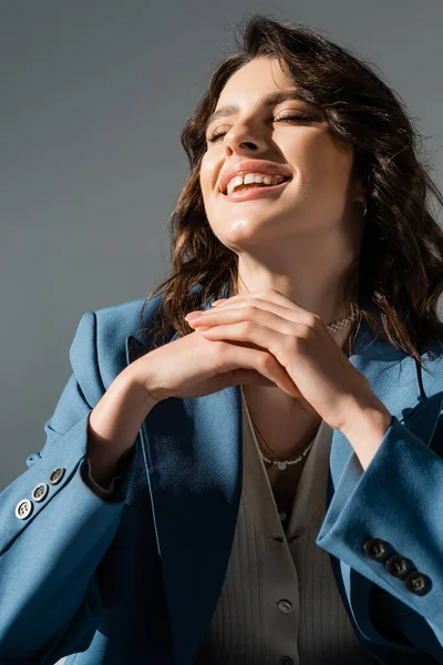Excited brunette woman in blue and fashionable blazer laughing with closed eyes isolated on grey — Stock Photo