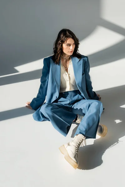 Full length of trendy woman in blue suit and white boots sitting on grey background with lighting and shadows — Stock Photo