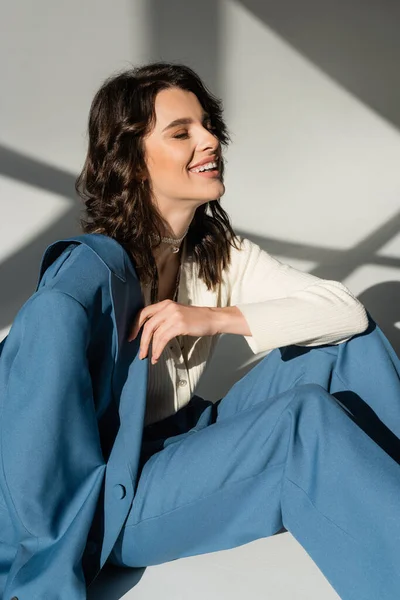 Happy brunette woman in blue suit laughing with closed eyes on grey background with lighting and shadows — Stock Photo
