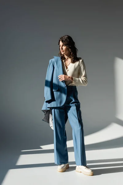 Full length of trendy woman in white boots standing with blue jacket on grey background with lighting and shadows — Stock Photo