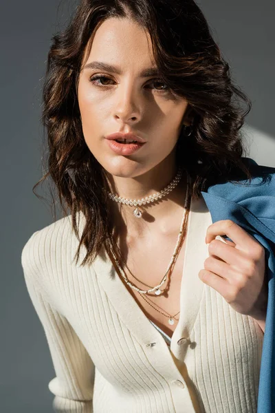 Brunette woman in white jumper and necklaces holding blue jacket and looking at camera on grey background — Stock Photo