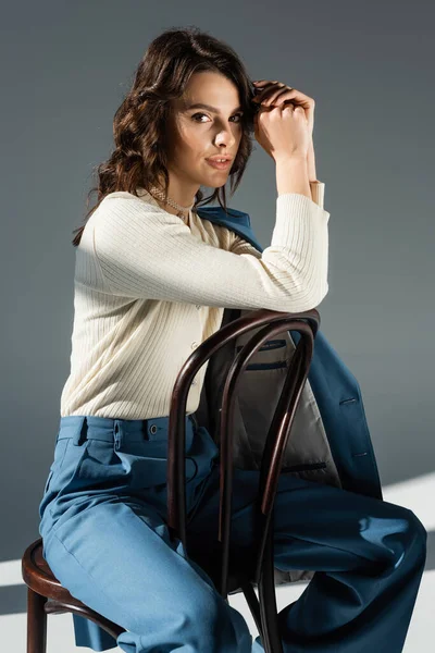 Donna positiva in maglione bianco e pantaloni blu seduta sulla sedia e che guarda la macchina fotografica su sfondo grigio — Stock Photo