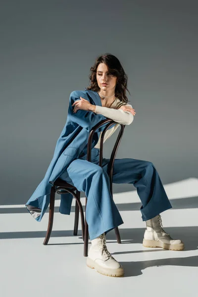 Longitud completa de la mujer de moda en pantalones y botas atadas posando en la silla cerca de chaqueta azul en gris - foto de stock