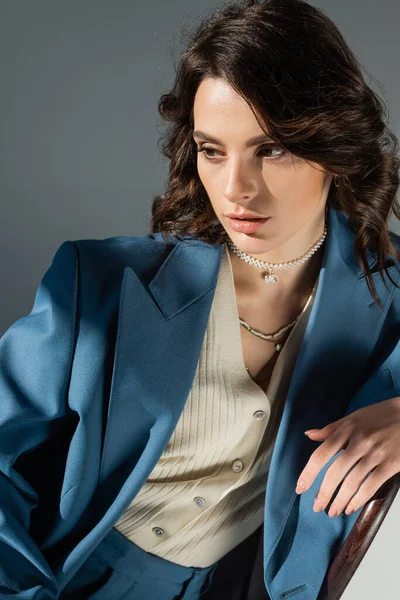Brunette woman in blue blazer and necklaces sitting and looking away on grey background — Stock Photo