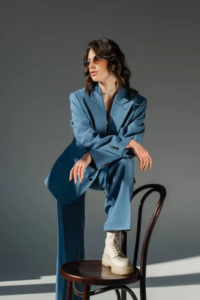 Brunette woman in blue fashionable suit stepping on chair and looking away on grey background — Stock Photo