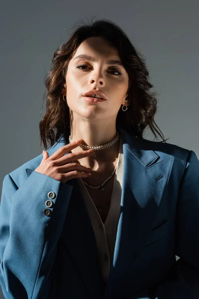 Sensual mujer morena en chaqueta azul tocando collar de perlas y mirando a la cámara aislada en gris - foto de stock