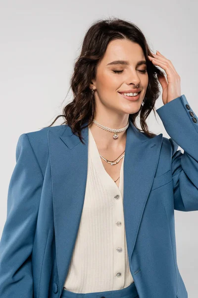 Pleased woman in blue blazer and necklaces posing with closed eyes and touching hair isolated on grey — Stock Photo