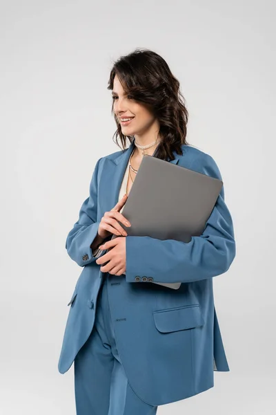 Mujer feliz en blazer azul de pie con el ordenador portátil y mirando hacia otro lado aislado en gris - foto de stock