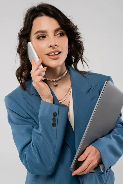 Donna positiva in blazer blu che parla su smartphone e tiene isolato il laptop su grigio — Foto stock