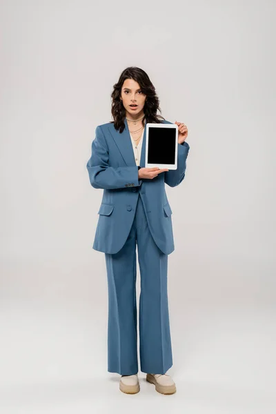 Piena lunghezza della donna sorpresa in abito blu mostrando tablet digitale con schermo bianco sul grigio — Foto stock
