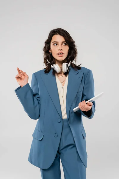 Trendy brunette woman with wireless headphones and digital tablet pointing with finger and looking away isolated on grey — Stock Photo